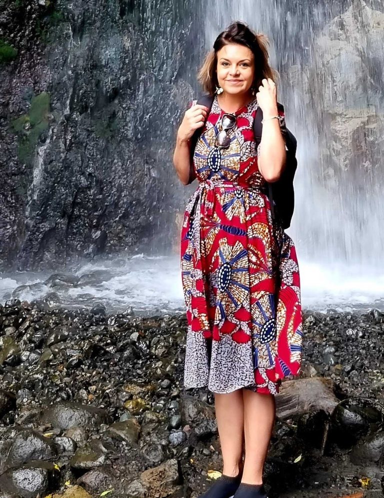 stephanie-safari-tanzanie-chute-eau Femme souriante en robe traditionnelle Tanzanie devant la chute d'eau Arusha, portant un sac à dos.